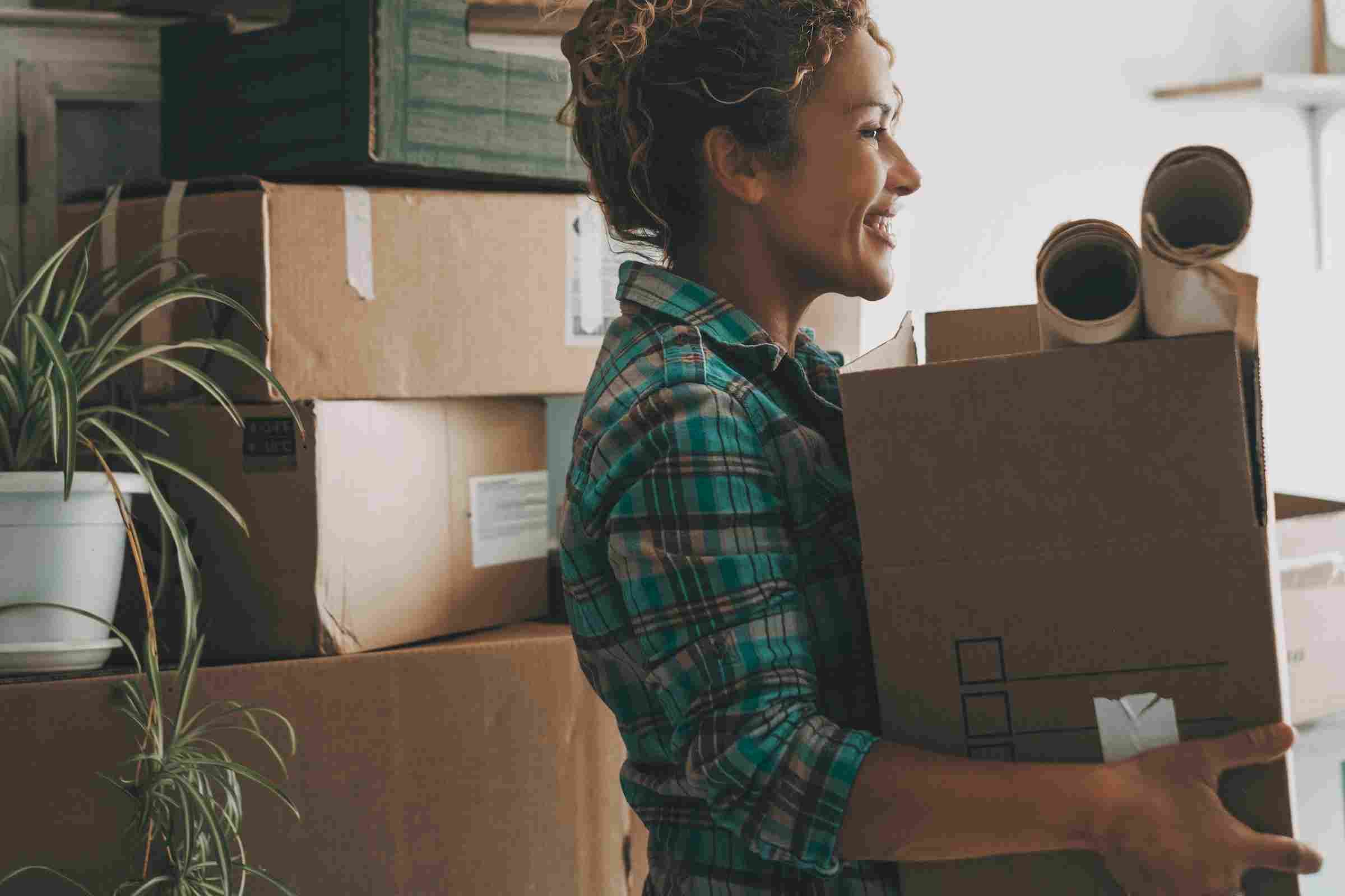 Sierra on 66 Smiling woman holding boxes and packing supplies, surrounded by moving boxes and a potted plant.