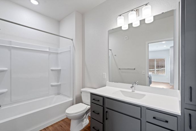 Sierra on 66 Modern bathroom with a bathtub-shower combo, toilet, gray vanity, and large mirror under three lights.