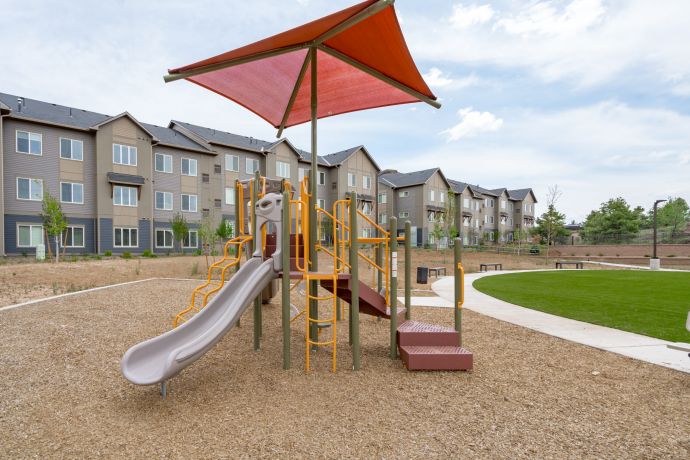 Sierra on 66 Playground with a slide and climbing equipment under a shade, in front of Sierra on 66 apartment buildings.
